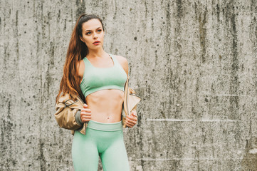 Outdoor portrait of young beautiful fit woman, wearing green activewear, athlete model posing next to grey urban wall background, sport fashion