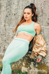 Outdoor portrait of young beautiful fit woman, wearing green activewear, athlete model posing next to grey urban background, leaning on the wall, sport fashion