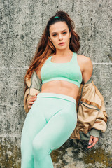 Outdoor portrait of young beautiful fit woman, wearing green activewear, athlete model posing next to grey urban background, leaning on the wall, sport fashion