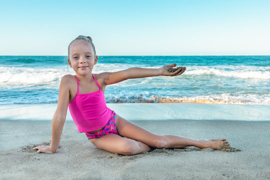 A playful cute little girl in a colorful pink swimsuit sitting on the sand, waves on the beach, outstretched hand showing horizon, ocean background, copy space