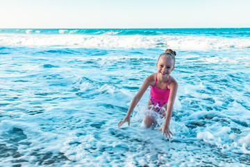A playful cute little girl in a colorful pink swimsuit sitting on the sand, waves on the beach, ocean background, copy space