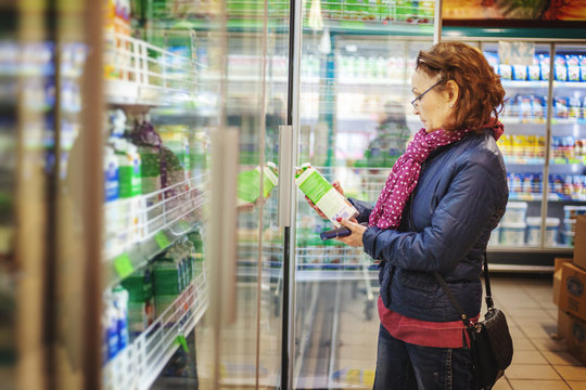 Mature Woman 60 Years Old Pensioner Chooses A Pack Of Milk From A Shelf In A Grocery Supermarket, Reads The Label