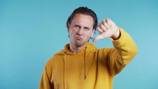 Young handsome man standing on blue studio background expressing discontent and showing thumb down gesture to camera. Portrait of guy with sign of dislike.