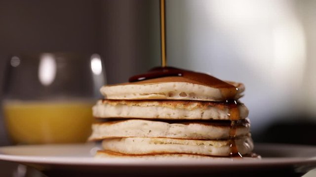 Maple syrup being poured on pancakes for breakfast with a glass of orange juice.