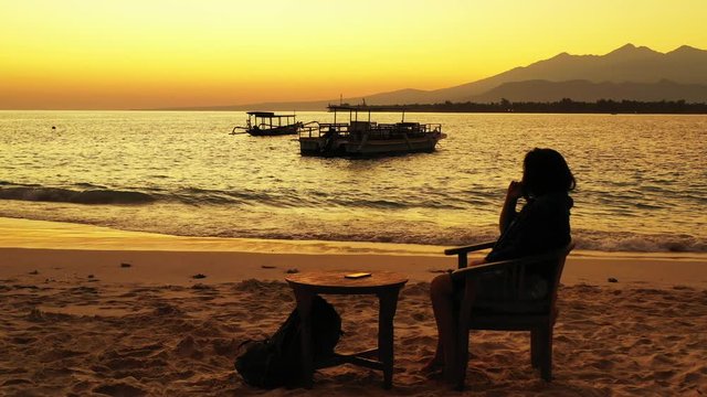 Woman Tourist Spending The Sunset Alone On The Beach Of Indonesia Sitting On A Chair With Backpack By The Table - Wide Pan Shot