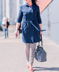 A woman in a blue coat, trousers and a large dark bag in her hands walks along a city street