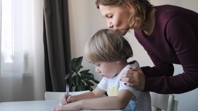 little boy sitting at the table at home and drawing, mother kissing and hugging him