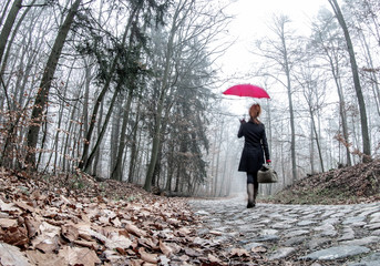 A woman with a red umbrella and bag is walking fast through a forest. It's on an old, narrow cobblestone street. Concept: loneliness