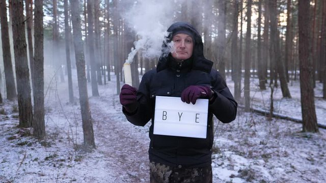 Man hold BYE sign and smoke plume burning out, look straight to camera then to empty chimney and walk away. Winter forest area, joker show lightbox with one word and slake smoke jet