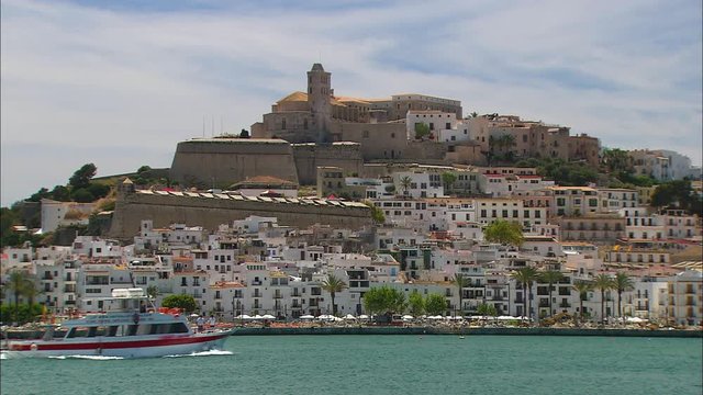 Medium low-angle still shot of beautiful white apartment buildings on a historical greek hilly island. At the hilltop is a historical museum with wind towers. A passenger ship sails along the island