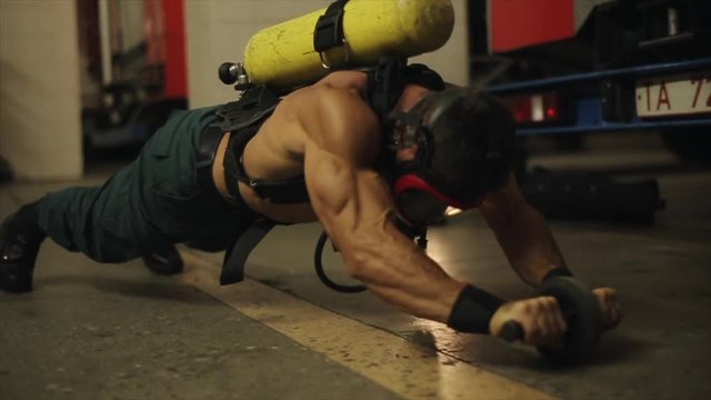 Half-naked fireman wearing mask and oxygen tank exercises with abs roller indoors