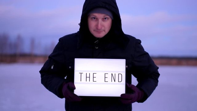 Positive man rise lightbox and show The End phrase, look straight to camera and down on words at board. Half length outdoor portrait of guy with white illuminated sign in hands, standing outdoor