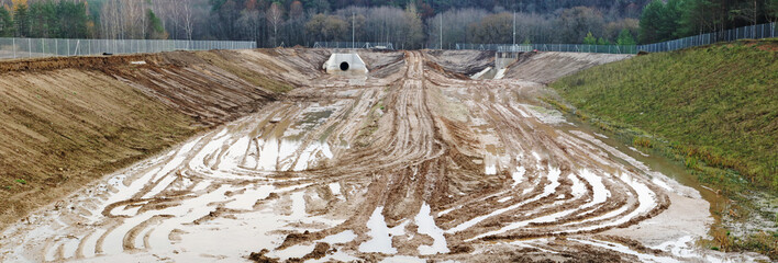 Large  concrete new collector for  sewer water  and heaps of wet sand with traces of heavy equipment wheels on a construction site in the forest panorama.