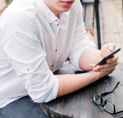 A young man in a white shirt and jeans sits at a table in a cafe, holds a phone in his hands and...
