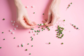 Women's hands make crafts from white and green beads. Pastel pink background. The concept of creativity, handwork, preparation for the holiday. Minimalism, top view, flat lay.