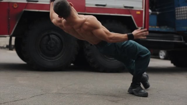 Slow motion. Attractive shirtless young athlete exercising push-ups with jumping. Big fire engine in the background. Side view