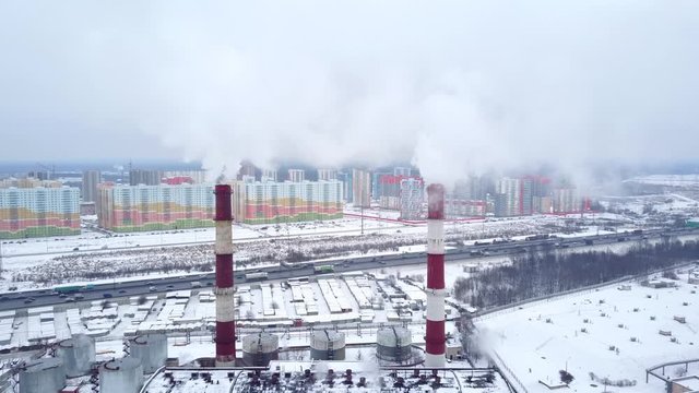 Pair of tall red-white chimney stacks at district heating station, aerial shot. Clouds of smoke curl out and fly away from tall tubes. Colored new residential houses seen on background
