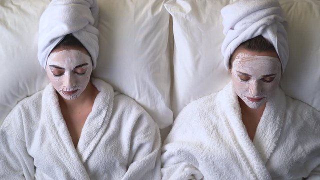 Two young women lying on bed with face mask on skin, enjoying beauty care in spa