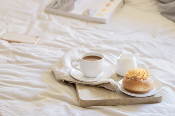 Fototapeta na wymiar Good morning scenery background. Breakfast In Bed, cup of coffee with sweet bun on wooden tray and book on beige white sheets.