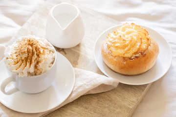 Morning Breakfast In Bed ,  cup of coffee  with foam milk and sweet bun on wooden tray on beige white sheets. Good morning scenery background. Scandinavian style.