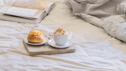 Good morning scenery background. Breakfast In Bed,  cup of coffee with sweet bun on wooden tray  and book on grey white sheets.