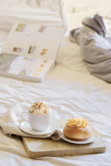 Fototapeta na wymiar Good morning scenery background. Breakfast In Bed, cup of coffee with sweet bun on wooden tray and book on grey white sheets.