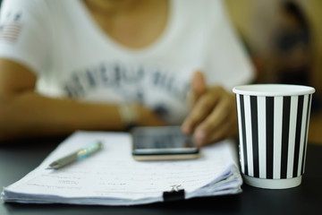 Hot coffee in paper cup with black and white stripes placed on the counter in front of a woman typing a job message on her mobile phone to send an email.