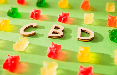Naklejka premium Wooden letters CBD on a green background and gelatin in the form of bears. View from above. The shadow of the sun falls on the background
