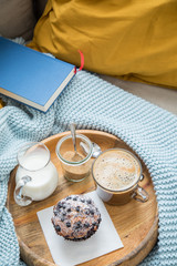 Blaubeermuffin mit Kaffee, Milch und Zucker auf Holztablett und gemütliches Sofa mit blau Wolldecke, gelb Kissen und Buch
