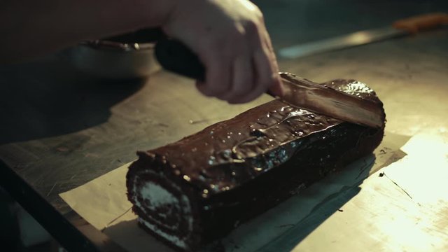 Hand with spatula is spreading chocolate on cocoa creamy swiss roll. Dessert lays on bakery parchment, stainless steel working table. Chef dabs soaked sponge cake, spreads melted chocolate evenly.