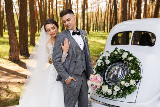 Elegant wedding couple, bride and groom embracing near white just married car with decoration of wreath with fresh flowers