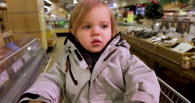 Curious blond haired toddler with blue eyes in casual warm clothing sitting in shopping cart and looking away at supermarket