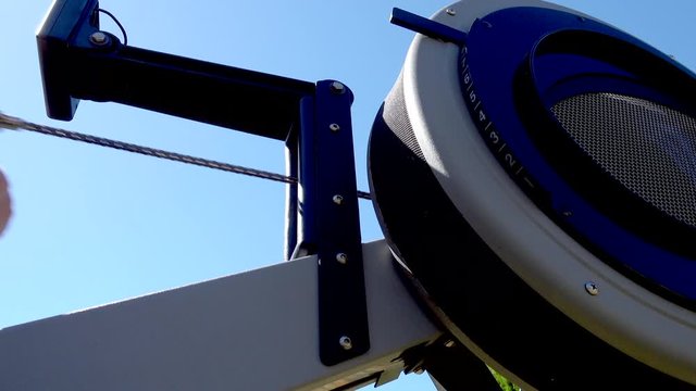 Closeup POV shot of a man pressing the display then gripping the handles to pull the chain on a sturdy, commercial rowing machine, which is being used outdoors, against a clear blue sky.