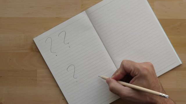 Man write many question marks on clean pocket-book page, top-down shot. Medium size notebook lie open on wooden table, man use pen to draw signs