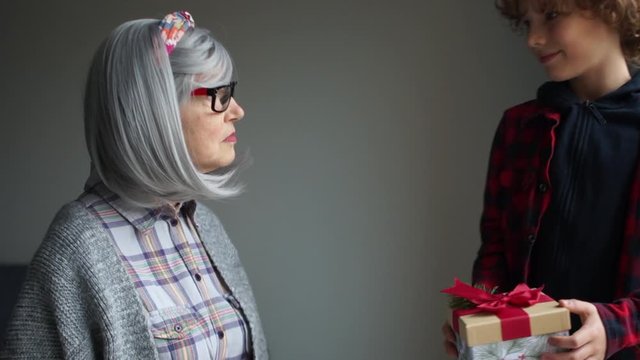Grandson teenager wishes Christmas to his grandmother. A woman in a gray wig and bright glasses. The boy is dressed in a checkered red-black shirt and holds a box with a gift