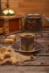 A Cup of coffee, coffee beans in a glass jar, brown sugar, coffee grinder on a wooden background.