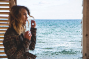 Caucasian girl stands near square window overlooking the sea from a wooden house. Traveling and nature concept. Unfocused percon.