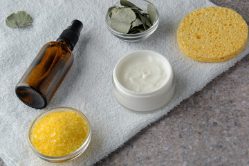 Fototapeta na wymiar Yellow salt, white cream, yellow sponge and dried eucalyptus leafs on white towel on grey stone table 