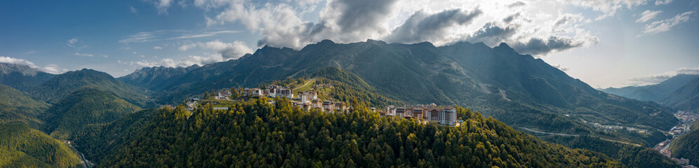 Fototapeta na wymiar Wide panorama of Rosa Khutor mounrain ski resort of Russia in sunny evening; olympic village surrounded by mountain ranges, valleys; summer winter widely known tourist destination for hiking, trekking