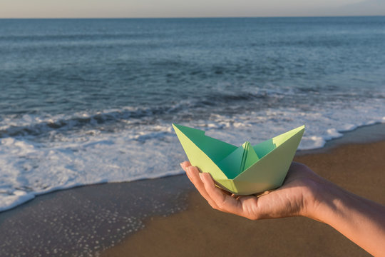 Closeup view of green paper boat in one single cupped hand of young white woman isolated on sunny early morning sea beach background. Horizontal color photography.