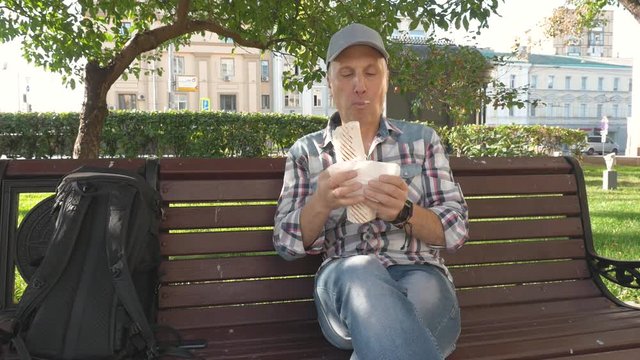 caucasian middle aged men gray plaid shirt blue jeans baseball cap Sitting in the Park on a bench Eating a big sandwich Wiping a napkin Focused face Summer Sunny day Greenery around Medium shot