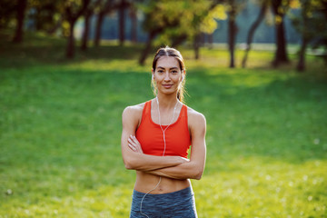 Fit healthy slim smiling caucasian brunette in sportswear and with ponytail standing on meadow with arms crossed. In ears are earphones. Sunny day in nature.