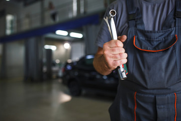 A mechanic in overalls with tools in his hands stands with his arms crossed on his chest. Work in the service station in the background.