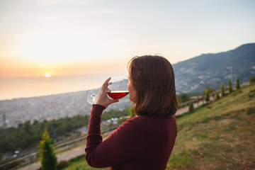 Woman drinking red wine on sunset mountains, close up of hand holding glass of wine. Elegant woman enjoying beautiful mountain and sea landscape on sunset.