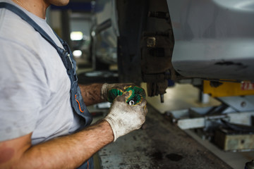 Car repair in the service station. Hands of a mechanic in overalls repairing the car on the lift without wheel, holding the tire and mechanical works.