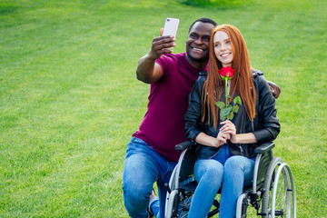 Fototapeta premium afro american man sitting on wheelchair ,his redhaired ginger girlfriend rolling stroller in autumn park. having romantic date.making a selfie on camera smartphone
