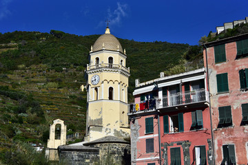 Fototapeta na wymiar Medieval church surrounded by colorful italian buildings and green agriculture terrasses of the Vernazza village of Cinque Terre under daylight. Italy 2019.