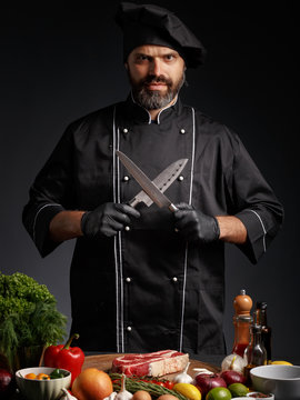 A male chef in black uniform sharpens knives near a table with groceries.