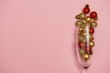 Champagne glass with golden and red baubles on pastel pink background, copy space. Minimal, New year concept. Top view, flat lay