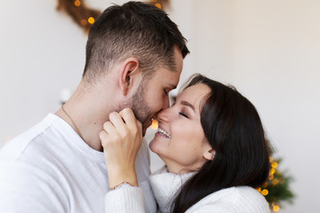 The concept of new year's love story. A guy and a girl in white cozy sweaters hugging, kissing, dancing and fooling around on the background of a white fireplace, decorative Christmas tree and bokeh.